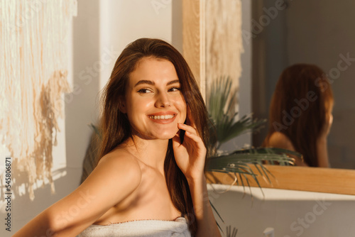 Happy excited brunette woman in white towel laughing in the bathroom. Attractive young lady is touching her face and smiling near the window.