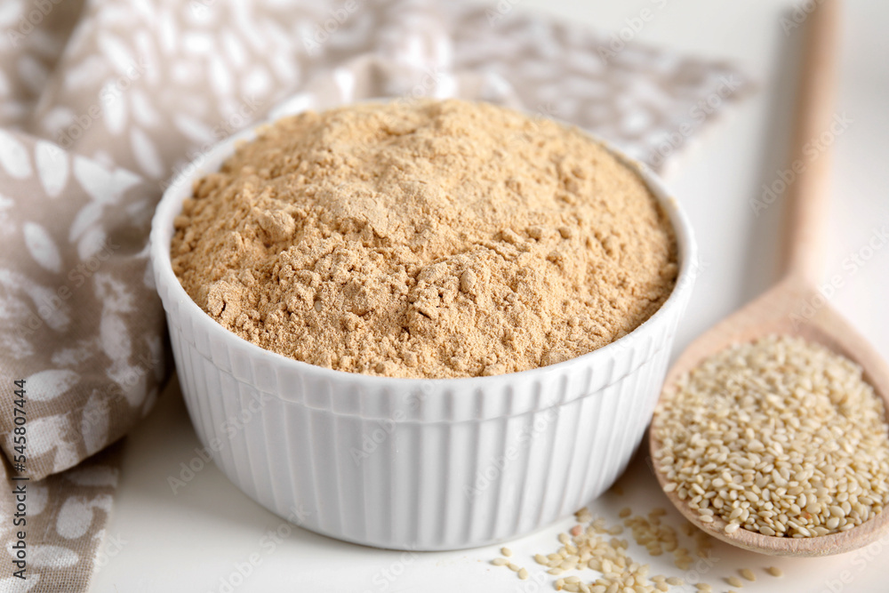 Sesame flour in bowl and wooden spoon of seeds on white table, closeup Sesame flour in bowl and wooden spoon of seeds on white table, closeup