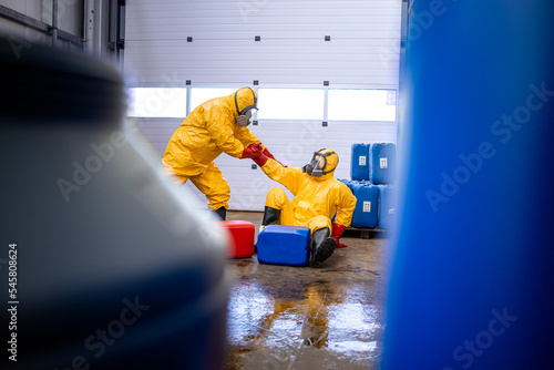 Accident in chemical factory, slippery floor, spilled chemicals. Worker in protection hazmat suit helping his colleague to stand up after the fall. photo