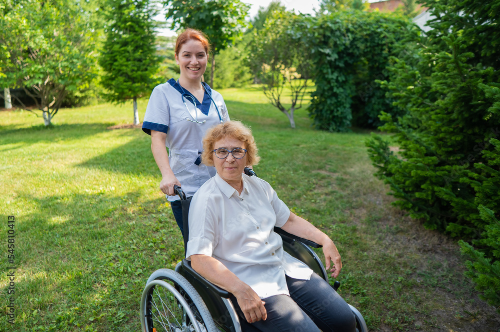 Caucasian female doctor walks with an elderly patient in a wheelchair in the park. Nurse accompanies an old woman on a walk outdoors. Caucasian female doctor walks with an elderly patient in a wheelchair in the park. Nurse accompanies an old woman on a walk outdoors.