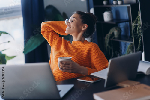 Foto Happy young woman holding coffee cup and keeping her feet on desk while sitting