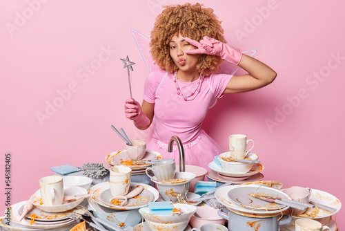 Lovely female fairy makes peace gesture over eye keeps lips rounded wears dress necklace and magic wand poses near sink and piles of dirty dishes around does house chores. Washing up concept photo