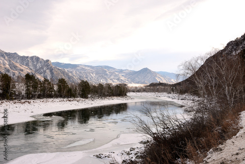 A view of a snow-covered valley with a flowing melted river on a winter evening. © Алексей Желтухин