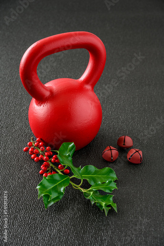 Holiday fitness, red kettlebell on a gym floor with a fresh sprig of festive Christmas holly and jingle bells
