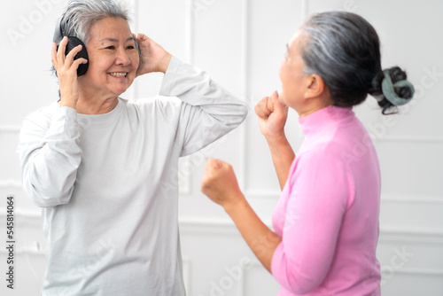 Senior women happy listening to music through earphones and singing song and smiling. Cheerful two female elderly have fun entertainment and relaxation at home after retirement. Pensioner lifestyle