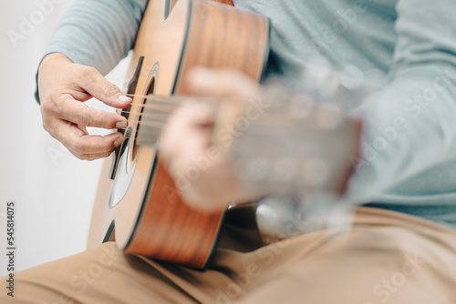 Senior man happy sitting playing acoustic guitar indoors as hobby after retirement. Elderly musician has fun singing a song with an instrument. Cheerful grandfather guitarist enjoys leisure activity.