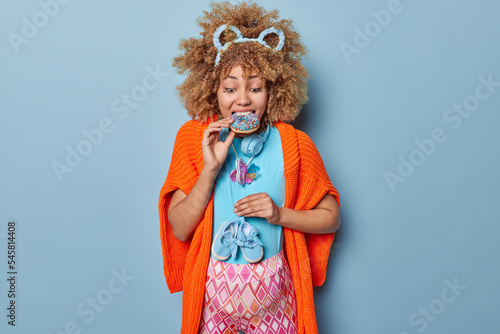 Hungry curly pregnant woman eats delicious glazed doghnut holds small baby shoes over tummy anticipates for baby going to become mother wears t shirt and leggings isolated over blue background photo
