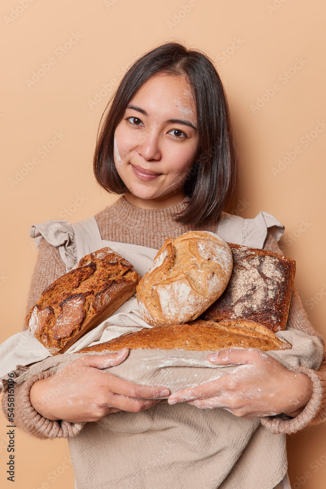 Asian female baker shop worker holds different loaves of bread baked in oven wears apron and knitted jumper looks directly at camera rests after baking isolated over beige background. Homemade food Asian female baker shop worker holds different loaves of bread baked in oven wears apron and knitted jumper looks directly at camera rests after baking isolated over beige background. Homemade food