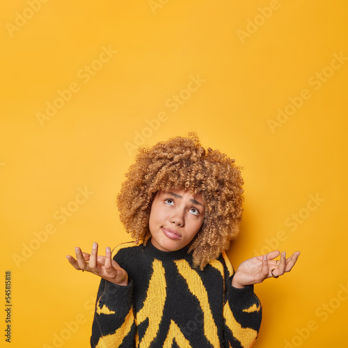 Fototapeta Unaware hesitant woman with curly hair spreads palms feels confused cannot decide on something wears casual jumper doesnt know what to do wears casual jumper isolated over yellow background