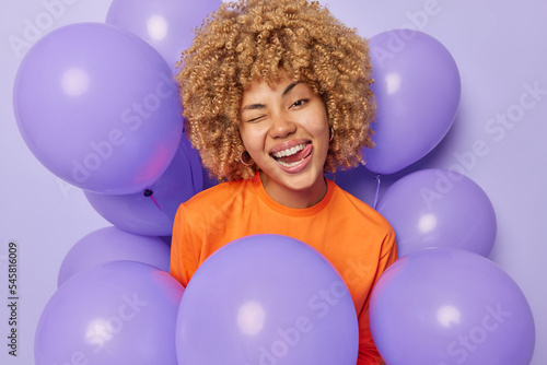 Funny playful woman with curly hair winks eye sticks out tongue enjoys festive event surrounded by inflated balloons comes on party to celebrate something wears orange jumper isolated on purple wall © wayhome.studio 