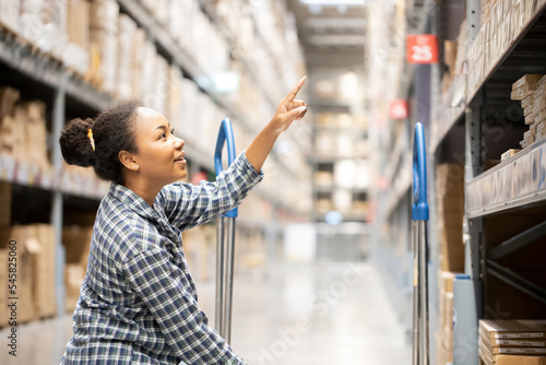 Portrait of young attractive African American woman auditor or trainee staff work looking up stocktaking inventory in warehouse store by computer tablet and stylus near products shelf. photo