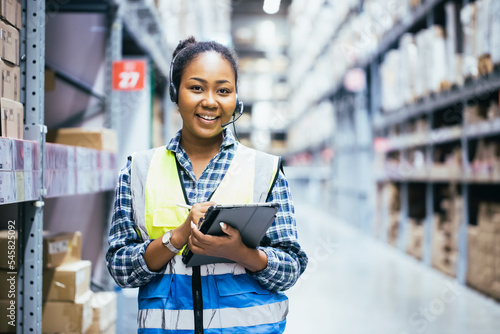 Portrait of young attractive African American woman auditor or trainee staff work looking up stocktaking inventory in warehouse store by computer tablet and headphones near products shelf. Call center