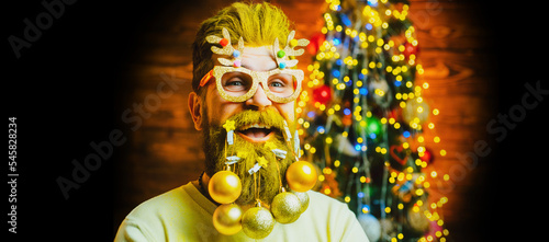 Concept of santa man with toy balls in long beard, x-mas decorations. Funny Santa man posing on vintage wooden background. Bearded modern Santa Claus decorating beard.