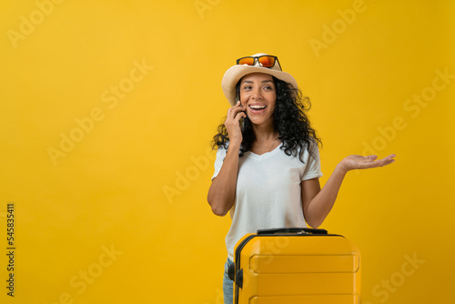 Happy Traveler curly latin woman wears white t-shirt with suitcase bag talking on phone isolated on yellow background.