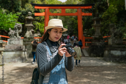 Asian Chinese woman photographer looking at her slr camera and checking photos near red torii gate entrance while visiting kasuga Taisha Shinto shrine in nara japan