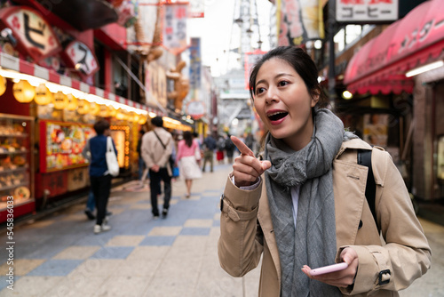 excited Asian Japanese female traveler finding out the right way to go and pointing at distance while using gps on the phone at nostalgic Shinsekai food street in Osaka Japan