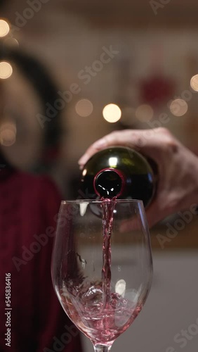 Vertical Screen: selective focus of red wine poured into glass from bottle by a man and a smiling woman looking with anticipation on blurred background while dating at home