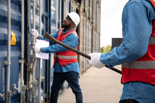 African American engineer holds a crowbar to open the door of a container chained to his assistant's hoof. Professional man in uniform and protective gloves standing in front of container.