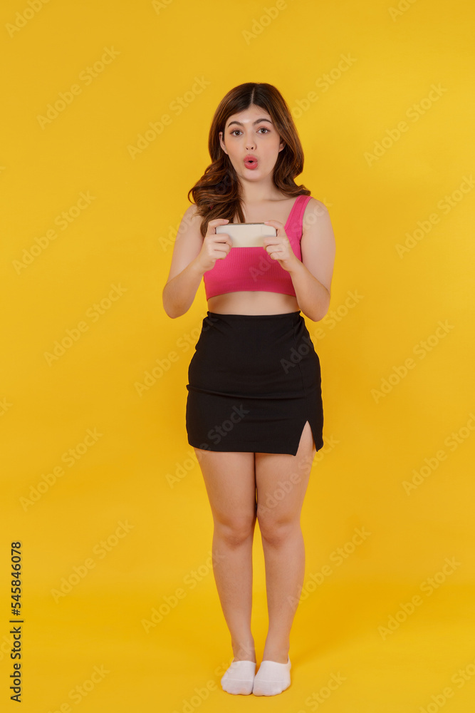 Portrait of Surprised young woman using mobile phone in her hands while standing isolated over yellow background Portrait of Surprised young woman using mobile phone in her hands while standing isolated over yellow background