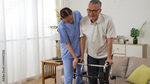 asian senior stroke patient undergoing rehab exercise with a walker at home. the woman nursing aide assists him during home visit