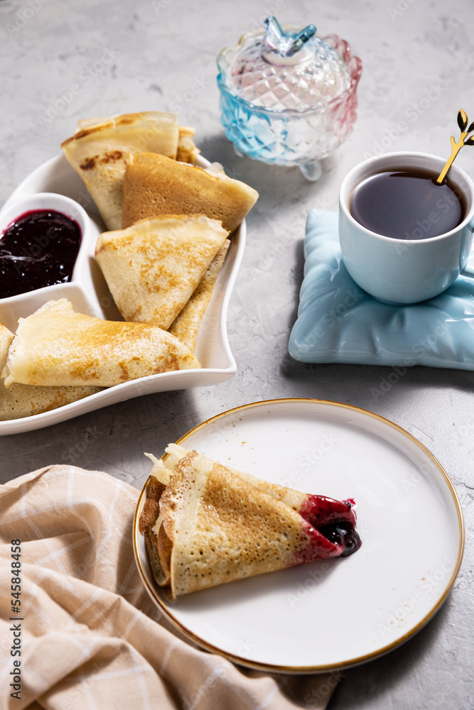 thin pancakes with jam in a heart-shaped plate with tea on a gray background thin pancakes with jam in a heart-shaped plate with tea on a gray background