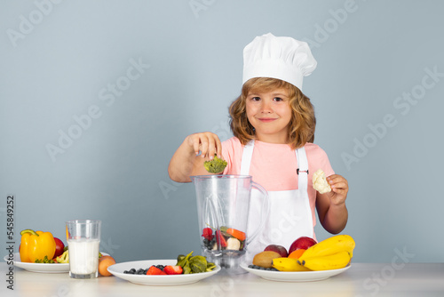 Child wearing cooker uniform and chef hat preparing vegetables on kitchen, studio portrait. Cooking, culinary and kids food concept.