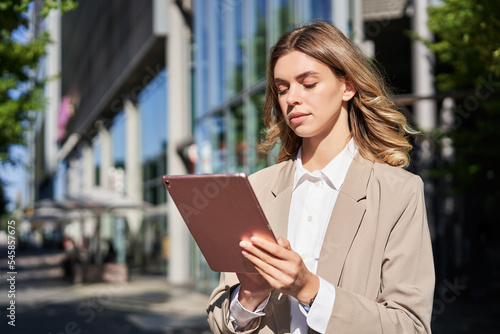 Portrait of corporate woman reads news, works on her digital tablet while on her way to office, stansd on street of city center in suit photo