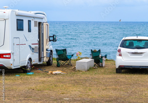 Motorhome parked with the sea in the background and two chairs with sea views. Tourist resting in the chair next to his dog on a leash. A seagull flies to the horizon.