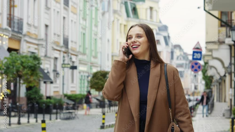 Cheerful brunette woman wearing casual brown coat walking street and talking on phone, blurred old building background. Tracking shot of young female in city. Concept of communication Cheerful brunette woman wearing casual brown coat walking street and talking on phone, blurred old building background. Tracking shot of young female in city. Concept of communication