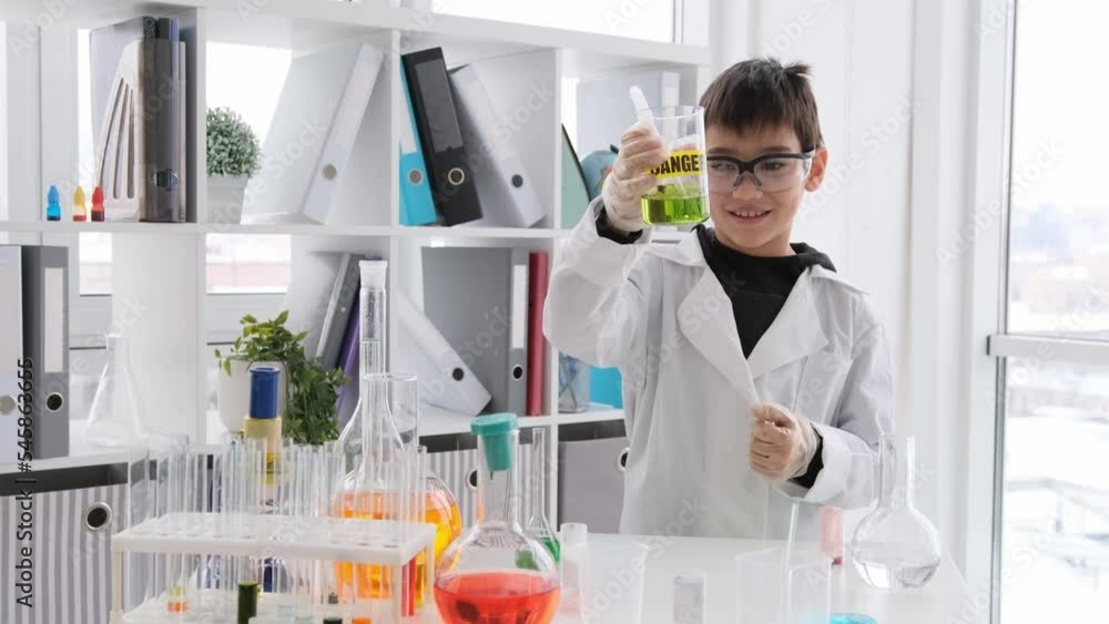 Schoolboy at chemistry lesson wearing protect glasses looking at bottle with green chemical liquid and mark danger. Pupil boy in lab studying and making experiments Schoolboy at chemistry lesson wearing protect glasses looking at bottle with green chemical liquid and mark danger. Pupil boy in lab studying and making experiments