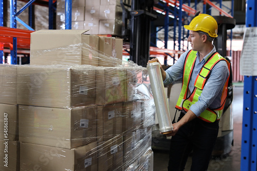 Caucasian warehouse worker man with hardhat and reflective jackets wrapping boxes in stretch film parcel on pallet while control stock and inventory in retail warehouse logistics distribution center