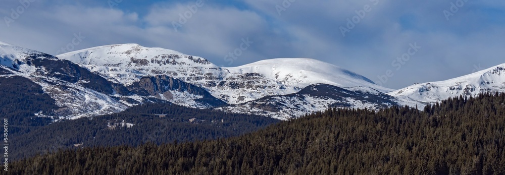 Beautiful winter panoramic natural mountain landscape. Attractive snowy peaks of Rila Mountains, Bulgaria. White clouds in dynamic blue sky, perfect conditions for tourism recreation and winter sports