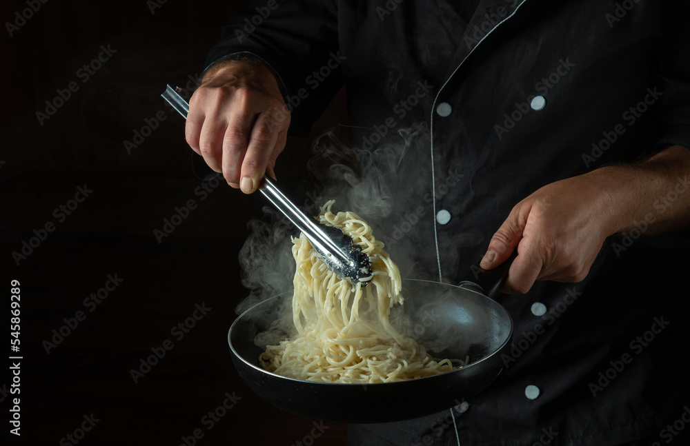 The cook fries spaghetti in a hot pan. Space for advertising on a black background. Italian national cuisine The cook fries spaghetti in a hot pan. Space for advertising on a black background. Italian national cuisine