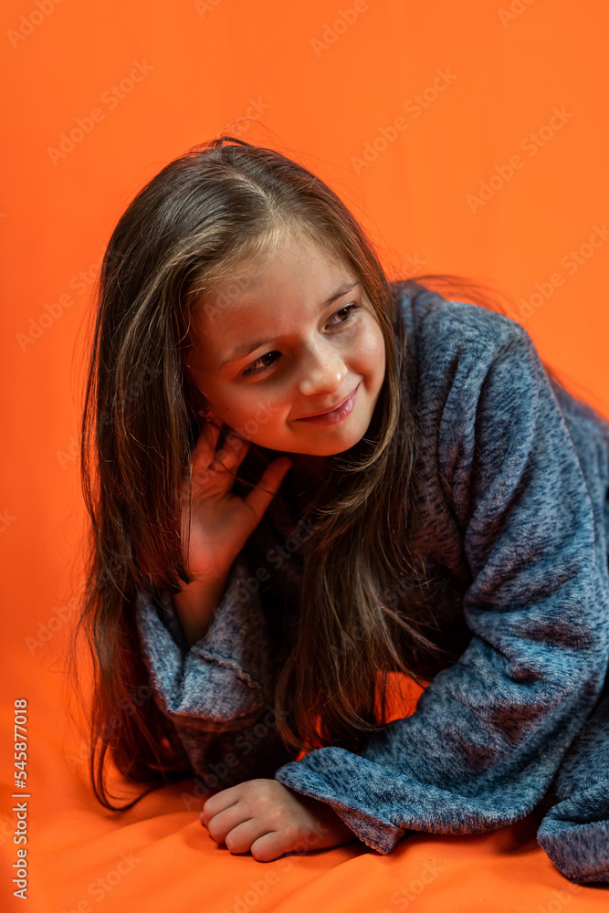 little girl with dark eyes in a gray robe is smiling while sitting on an orange background. little girl with dark eyes in a gray robe is smiling while sitting on an orange background.