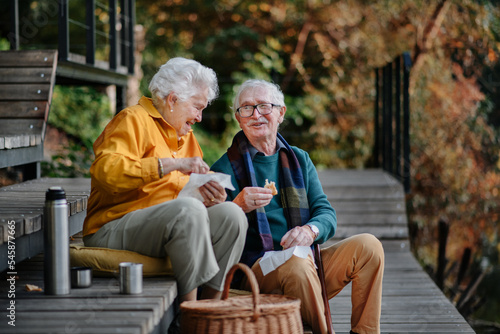 Happy senior couple in autumn clothes having picnic near lake after walk.