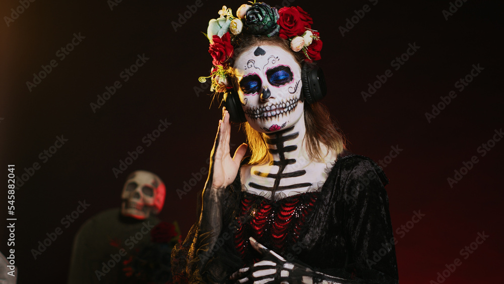Fototapeta premium Lady of death listening to music on headphones, wearing skull make up and santa muerte costume on day of the dead. Enjoying fun song on audio headset, celebrating mexican tradition. Handheld shot.