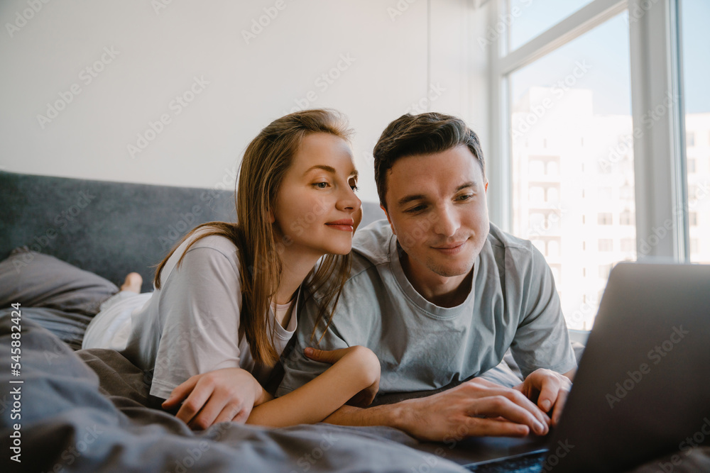 White young couple using laptop while resting on bed together White young couple using laptop while resting on bed together
