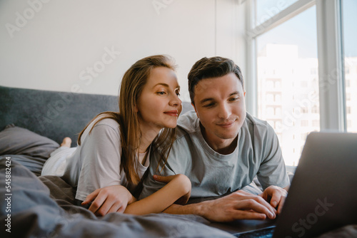 White young couple using laptop while resting on bed together