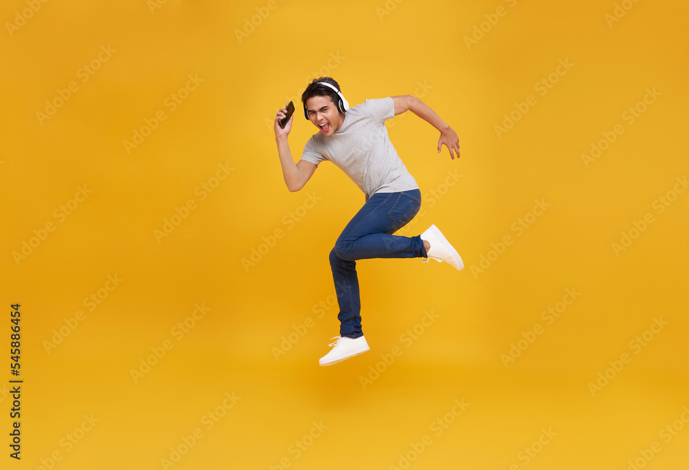Young handsome Asian man smiling holding smartphone and jumping wearing wireless headphone listening to music isolated over yellow background.