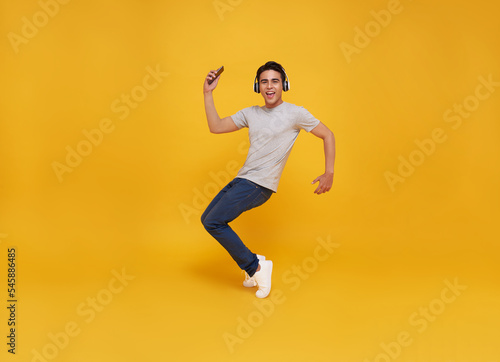 Young handsome Asian man smiling holding smartphone and jumping wearing wireless headphone listening to music isolated over yellow background. © NaMong Productions