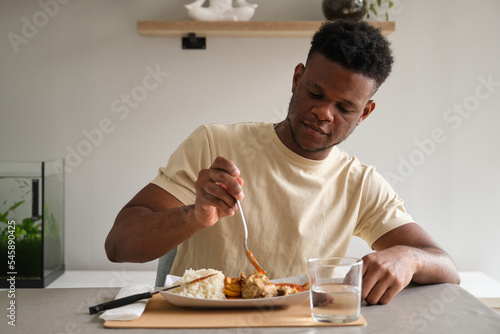 Fotografie Young african man eating fried chicken, rice and plantain with tomato sauce plate in the living room