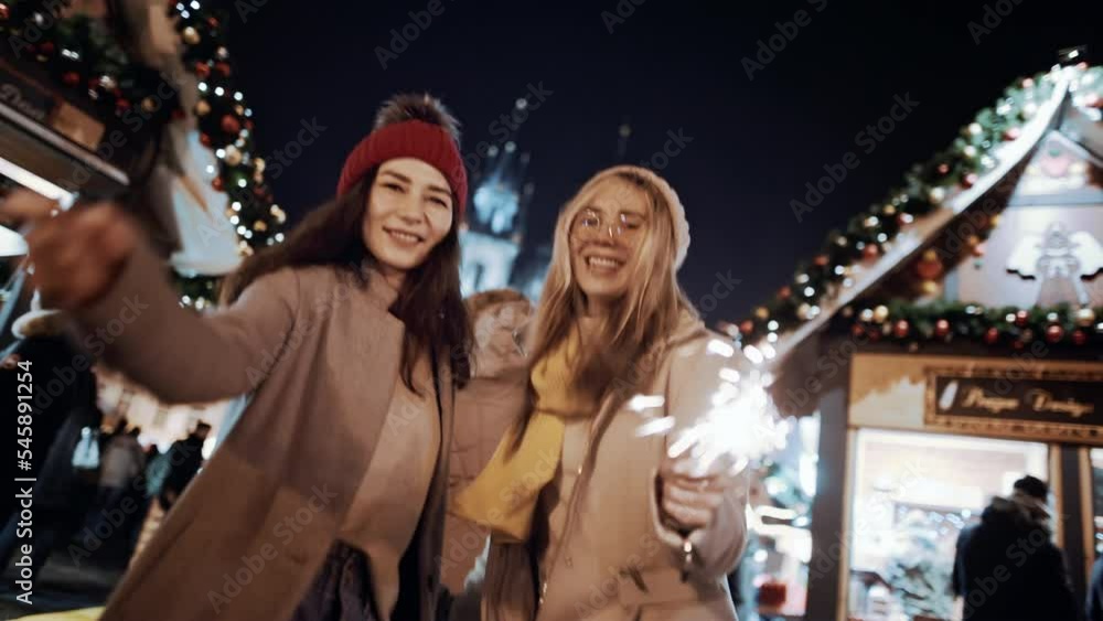 Celebrating New Year holidays vacation. Happy dancing female friends with sparklers smile at decorated town square. Xmas Christmas winter atmosphere. Excited girls people holding lights in hands. Celebrating New Year holidays vacation. Happy dancing female friends with sparklers smile at decorated town square. Xmas Christmas winter atmosphere. Excited girls people holding lights in hands.