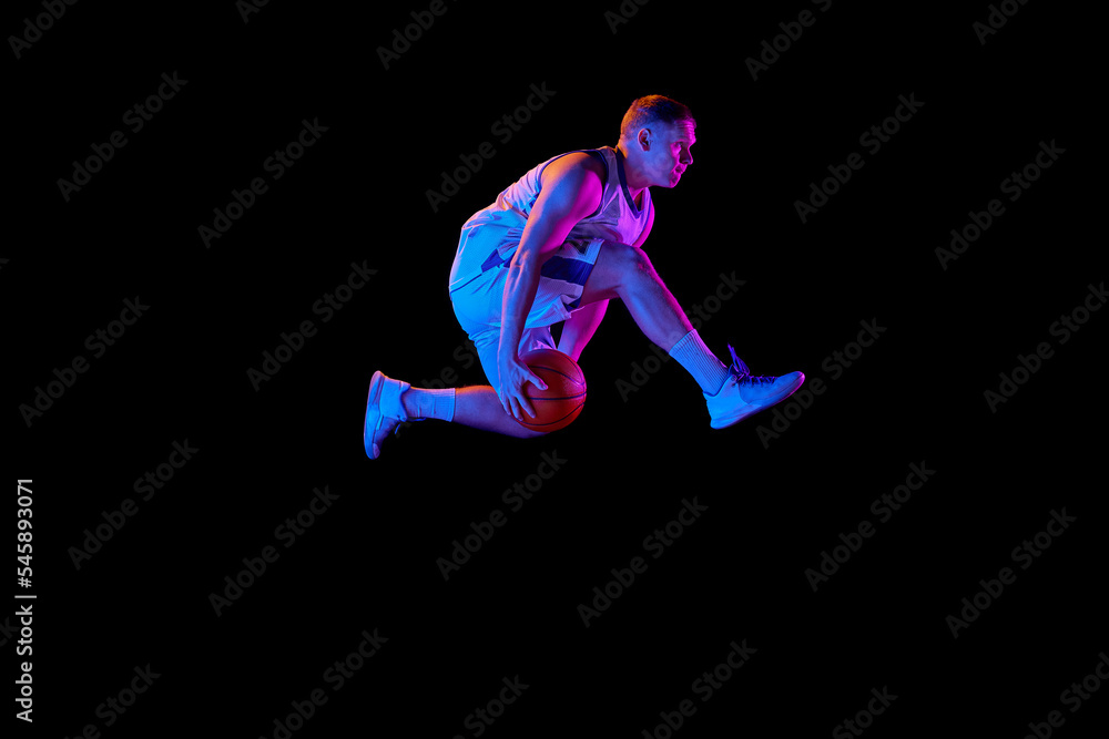 Active athletic male basketball player jumping with basketball ball isolated over dark background in purple neon light. Concept of energy, professional sport, hobby.