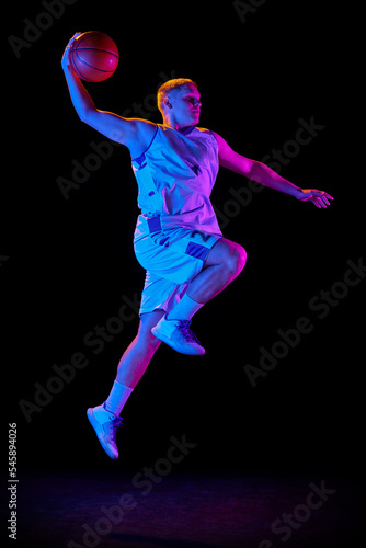 Active athletic male basketball player jumping with basketball ball isolated over dark background in purple neon light. Concept of energy, professional sport, hobby. © master1305