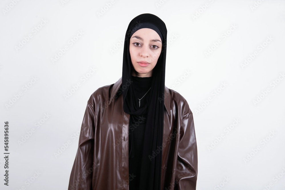 Joyful young beautiful muslim woman wearing hijab and leather jacket over white background looking to the camera, thinking about something. Both arms down, neutral facial expression.