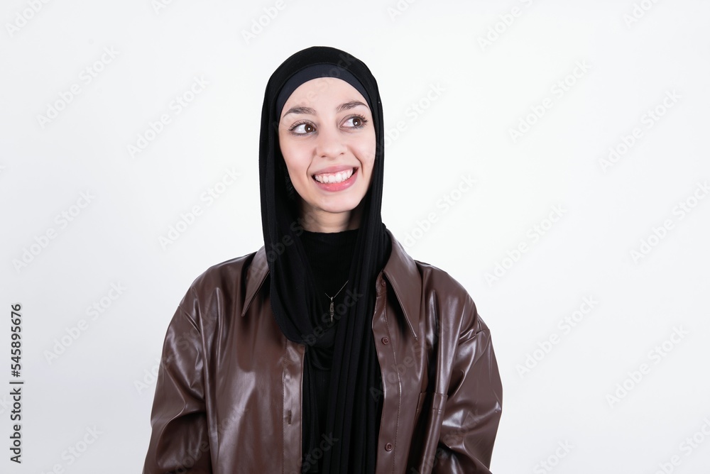 Oops! Portrait of young beautiful muslim woman wearing hijab and leather jacket over white background  clenches teeth and looks confusedly aside, realizes her bad mistake,