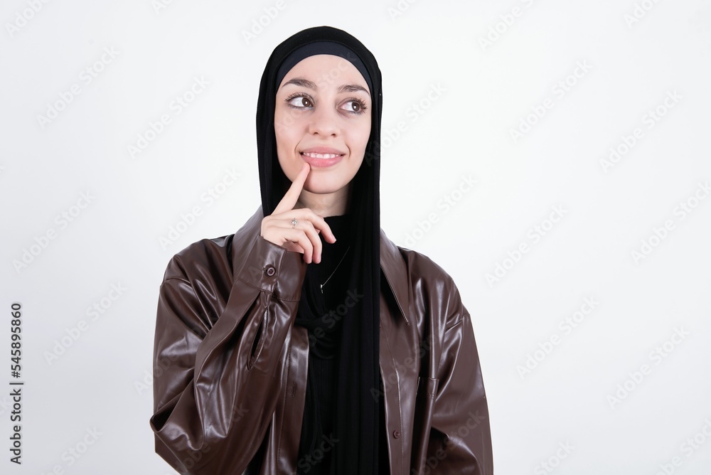 young beautiful muslim woman wearing hijab and leather jacket over white wall with thoughtful expression, looks to the camera, keeps hand near face, bitting a finger thinks about something pleasant. young beautiful muslim woman wearing hijab and leather jacket over white wall with thoughtful expression, looks to the camera, keeps hand near face, bitting a finger thinks about something pleasant.