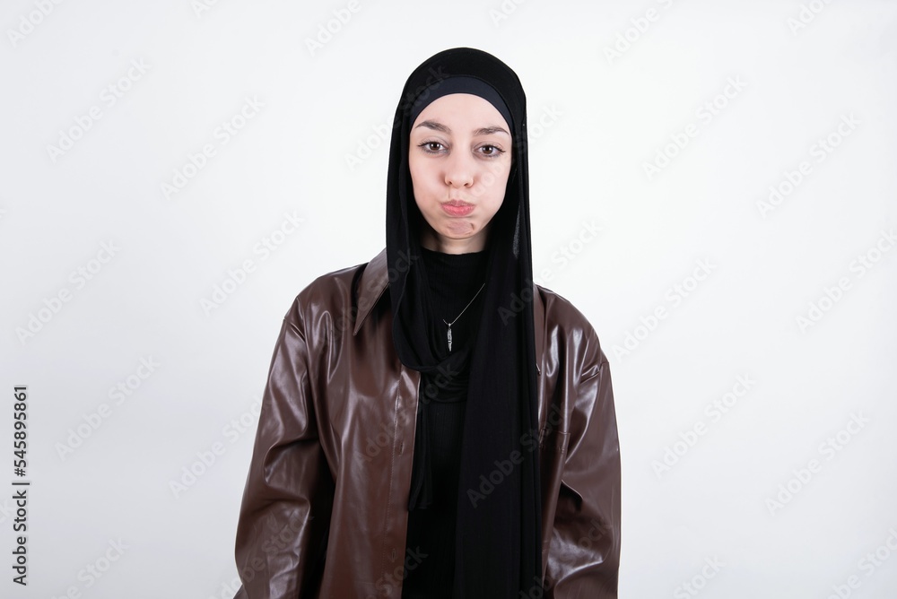 young beautiful muslim woman wearing hijab and leather jacket over white background puffing cheeks with funny face. Mouth inflated with air, crazy expression.