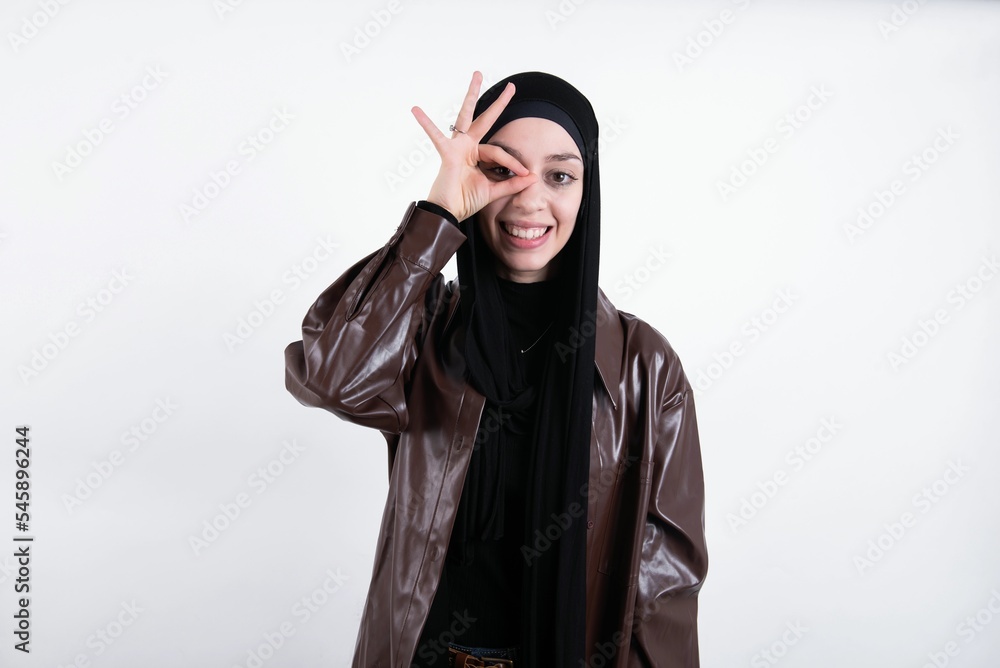 young beautiful muslim woman wearing hijab and leather jacket over white background with happy face smiling doing ok sign with hand on eye looking through finger.