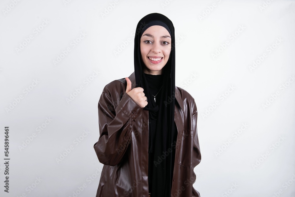young beautiful muslim woman wearing hijab and leather jacket over white background giving thumb up gesture, good Job! Positive human emotion facial expression body language. young beautiful muslim woman wearing hijab and leather jacket over white background giving thumb up gesture, good Job! Positive human emotion facial expression body language.